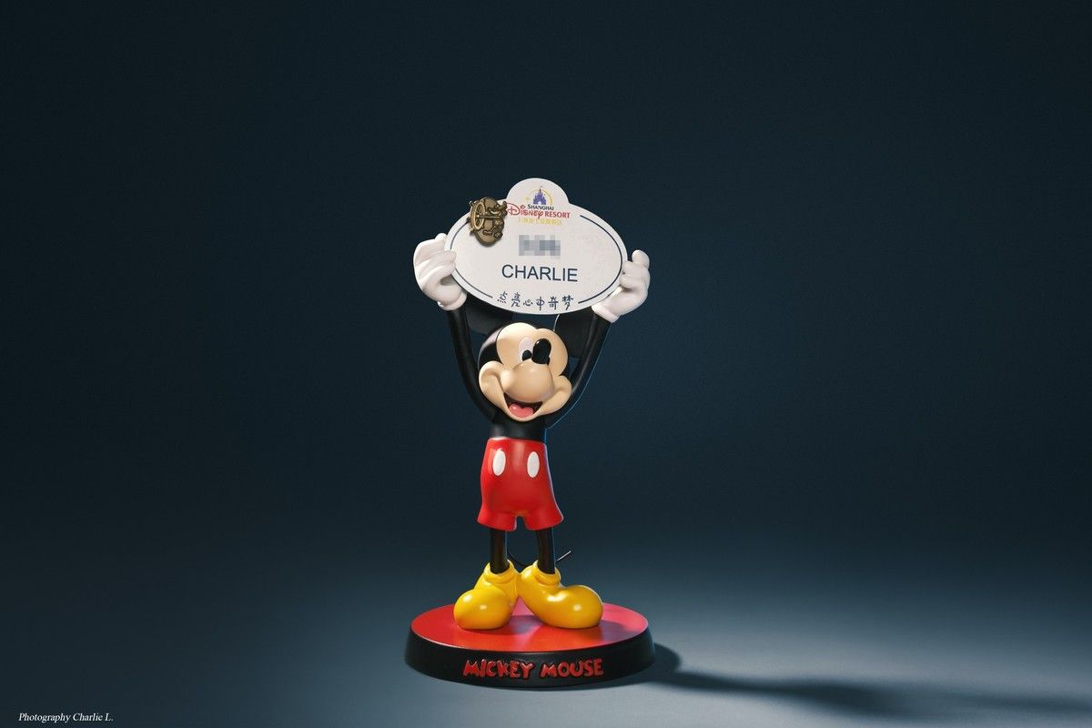 A Shanghai Disney Resort cast member name tag, reading CHARLIE, held up by a Mickey Mouse figure. Photographed by Charlie Liu.